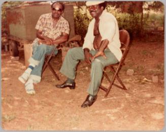 1982 [Two men sitting outside in brown folding chairs, man on left in sunglasses and man on right has shirt unbuttoned]