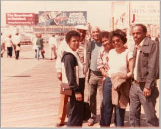[Three woman and three men posed together on wood boardwalk, billboards and gift shop signs around them]