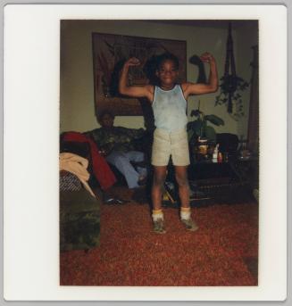 [Boy in tank top posing to flex arm muscles, a person is sitting on the couch behind him]