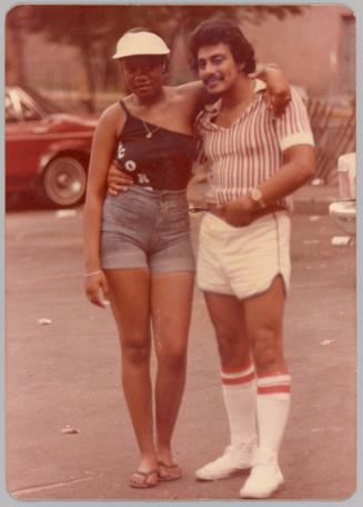 [Woman and man with arms around each other in parking lot, dressed in summer clothing, man wearing knee high socks]
