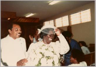 [Woman in wedding dress smiling and holding veil with man in white on her left]
