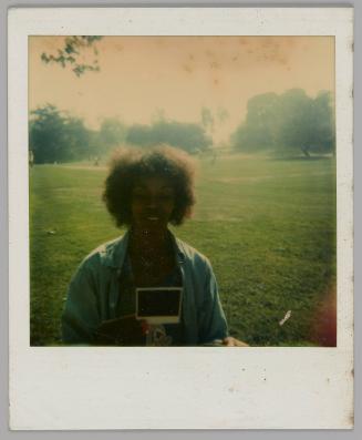 [Woman holding and looking down at polaroid in a park with a large empty field, she is wearing an unbuttoned blue shirt]