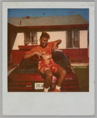 [Woman in tie dye overall shorts and pink t-shirt sitting on trunk of red car, car parked by bungalow with barred windows]