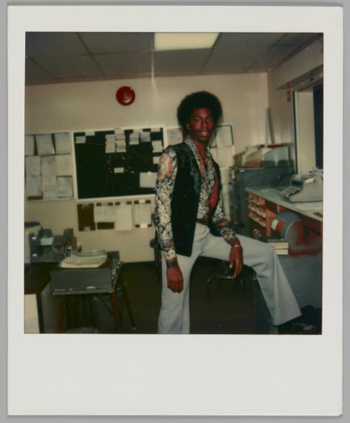 [Man in flared pants, patterned shirt and open black vest standing with one leg propped up in a classroom, red fire alarm on wall behind him]