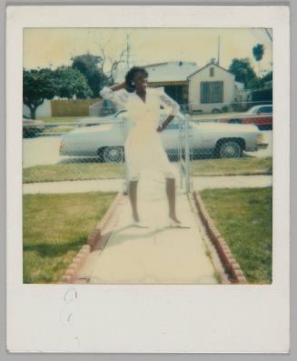 [Woman in white dress and lace sleeves posing with one hand on hip and one hand behind her head , she is standing on walkway of front lawn]