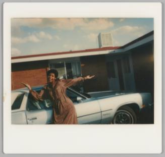 [Woman in dress posing with her arms spread wide in front of a parked car, one storey building behind her]