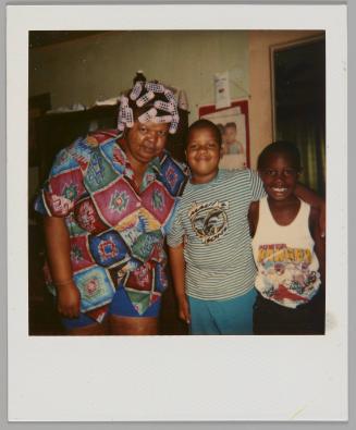 [Woman with pink curlers in her hair and wearing a colourful short-sleeved shirt standing with two boys, boy on right is wearing a Power Rangers tank top]