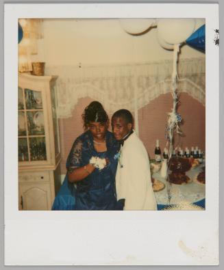 [Young woman in dress, black lace shrug, and white corsage beside young man in white suit jacket, behind them are balloons, drinks, and food]