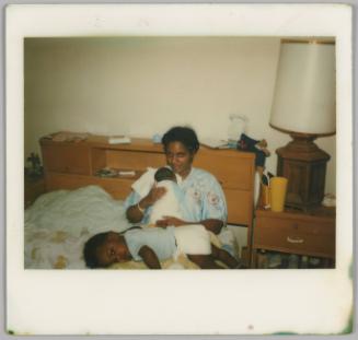 [Woman in bed holding sleeping baby against her chest, a toddler in a diaper is lying across the woman's lap, the bed's headboard has built in shelves]