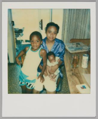 [Woman in shiny top sitting with baby on her lap, boy in white tank top and blue shorts standing beside her with his hand on her knee]