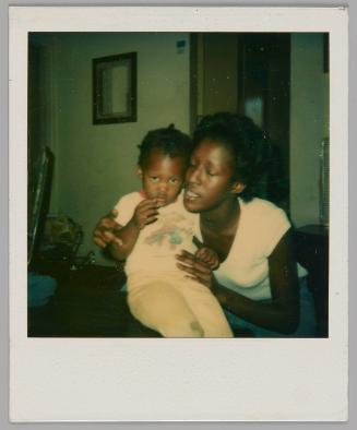 [Woman in white t-shirt sitting and holding a child on her lap, child is looking at the camera and touching their mouth]