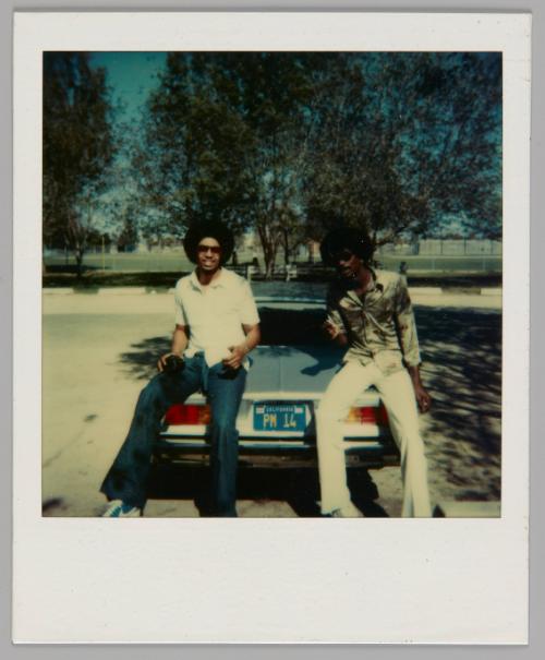 [Two men sitting on the trunk of a car in parking lot surrounded by trees, the license plate is "PM 14"]