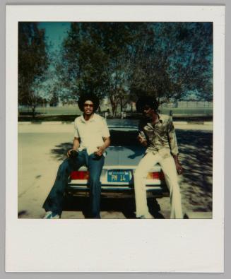 [Two men sitting on the trunk of a car in parking lot surrounded by trees, the license plate is "PM 14"]