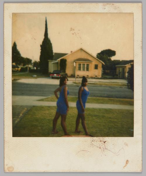 [Two women in tight dresses posing on grass lawn by a sidewalk, across the street is a yellow house]