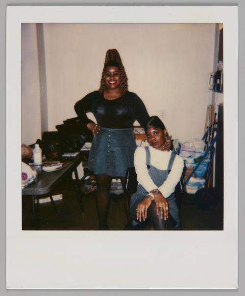 [Woman in black shirt and denim skirt standing with one hand on her hip and other hand on the woman sitting down beisde her, woman sitting has her hands crossed over her knees]