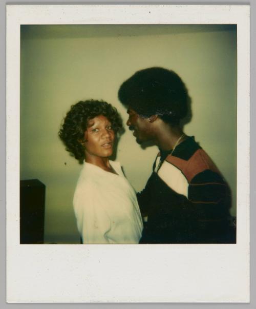 [Woman with short curly hair and a white shirt looking towards camera, she is with a man in a collared shirt who is looking at her]