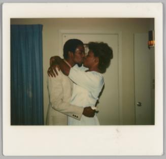 [Man in light-coloured suit kissing woman in white dress, they are inside and there is a blue curtain and door behind them]