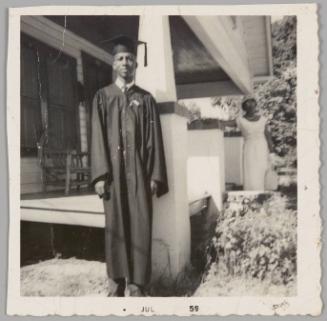 [Young man in cap and gown standing outside by front porch, woman in light-coloured dress standing behind him]