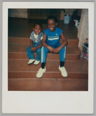 [Two kids sitting on steps, boy on right wearing blue t-shirt and track pants with white stripes and white velcro shoes]