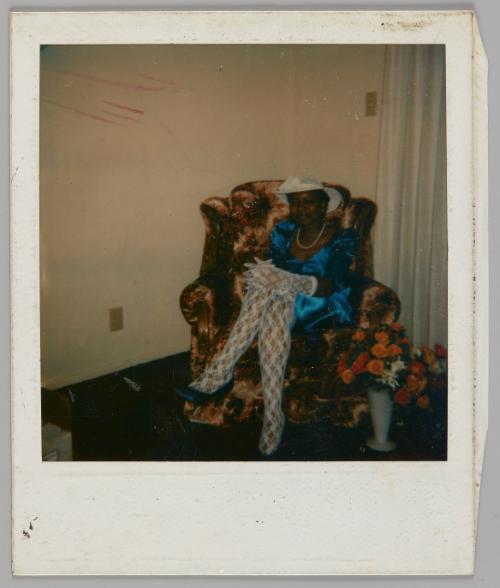 [Woman in white lace gloves and stockings with a white hat and blue dress sitting in armchair with a vase of flowers by her feet]