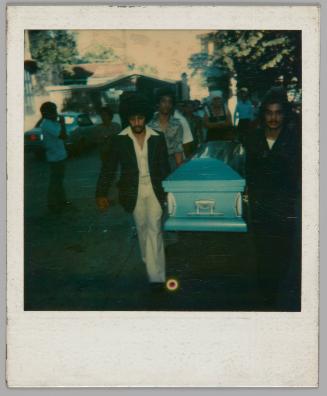 [Pallbearers carrying blue casket outside, man on left is wearing a dark jacket and a light shirt with a wide collar]