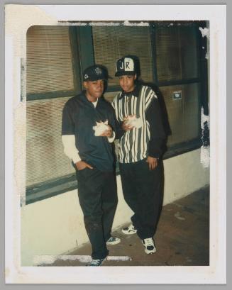 [Twp teenagers posing with one hand in pocket and one hand holding take-out food, person on right wearing white and black striped shirt and "JR" baseball cap]