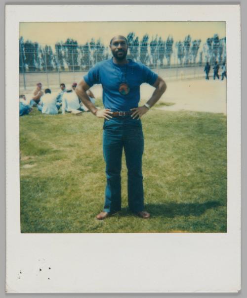 [Man in blue t-shirt, jeans and brown belt standing outside with his hands on his hips, circle of people sitting behind him]