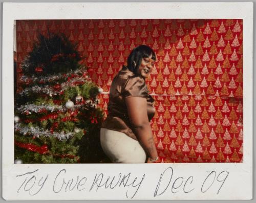 Toy Giveaway Dec 09 [Woman in shiny brown short-sleeved top looking back at camera, wall is covered in Christmas tree wrapping paper and there is a Christmas to the left]