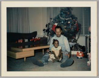 [Girl sitting on the floor between a man's legs with her doll, man is looking down at her, there is a Christmas tree behind them]