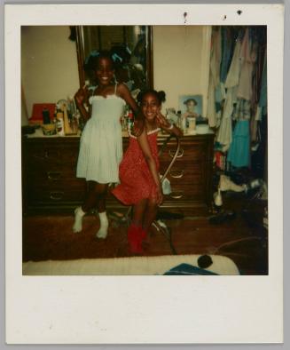 [Two girls, one on left wearing blue dress and one on right wearing red dress, standing in front of dresser beside a closet filled with clothes]