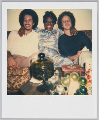 [Woman in patterned outfit sitting on couch between two young with arms wrapped around them]