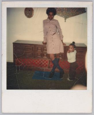 [Young woman posing against cabinet, toddler stands next to her]