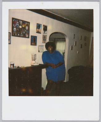 [Woman with afro and blue dress leaning against mantel near doorway]