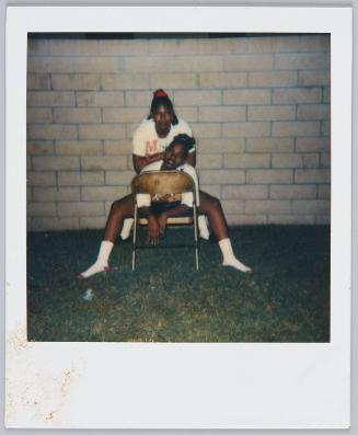 [Two young women posing in front of brick wall, one sits on a chair turned backwards]