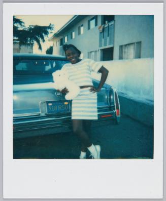 [Girl wearing blue and white stiped top and matching skirt leans against car and holding a white stuffer animal]