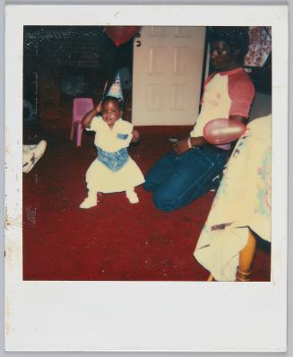 [Toddler girl wearing party hat and standing on red carpeted floor]