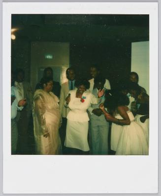 [Group gathered around woman in white outfit holding a microphone]