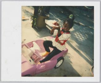 [Little girl sitting in pink toy car holding up a candied apple]