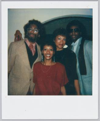 [Two women standing in between two men in suits, man on right wearing grey suit and sunglasses]