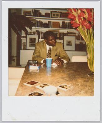 [Man in suit sitting at table, polaroid pictures scattered on table]