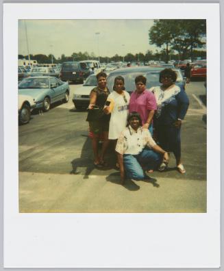 [Group of five woman posing in parking lot, two women on left holding polaroids]