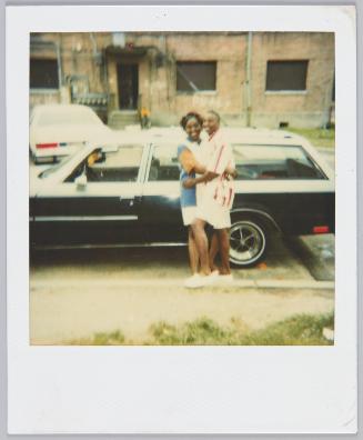 [Young couple hugging in front parked car, man on right wears red and white striped shirt]