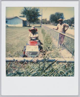 [Woman in hat sitting on lawn mower, man standing other side of gate next to her]