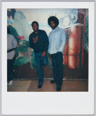[Two men standing in front of decorated wall, man on right wears blue button up and has an afro]