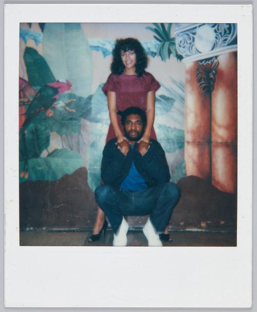 [Couple in front of decorated wall, man squatting and woman stands behind him as they hold hands]