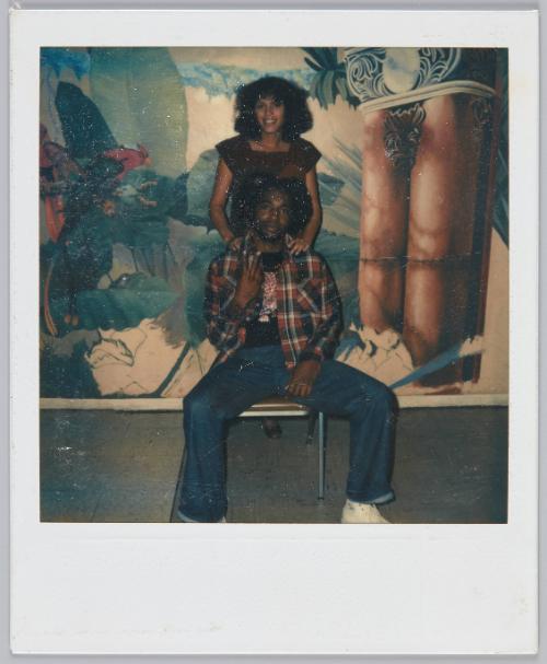 [Couple in front of decorated wall, man sits on chair and woman stands behind him]
