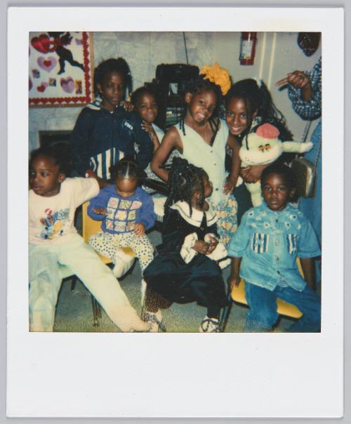 [Four children sitting on yellow chairs, four girls gathered around behind the seated children, girl standing on far right is holding a frog plush toy]