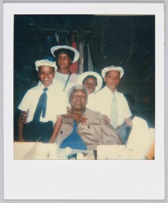 [Four children in white hats gathered around middle-aged man sitting in chair]