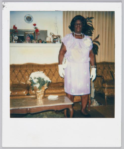[Woman in pink dress and white gloves standing in living room in front of golden yellow couch and armchair set, white flowers on coffee table beside her]