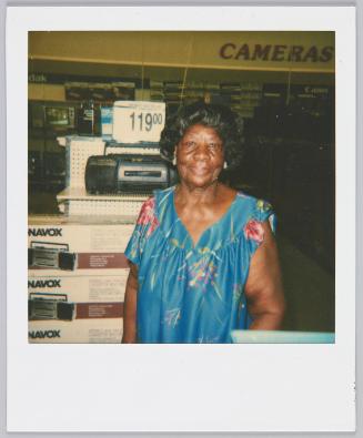 [Middle-aged woman standing in store in front of display of cameras and radios]